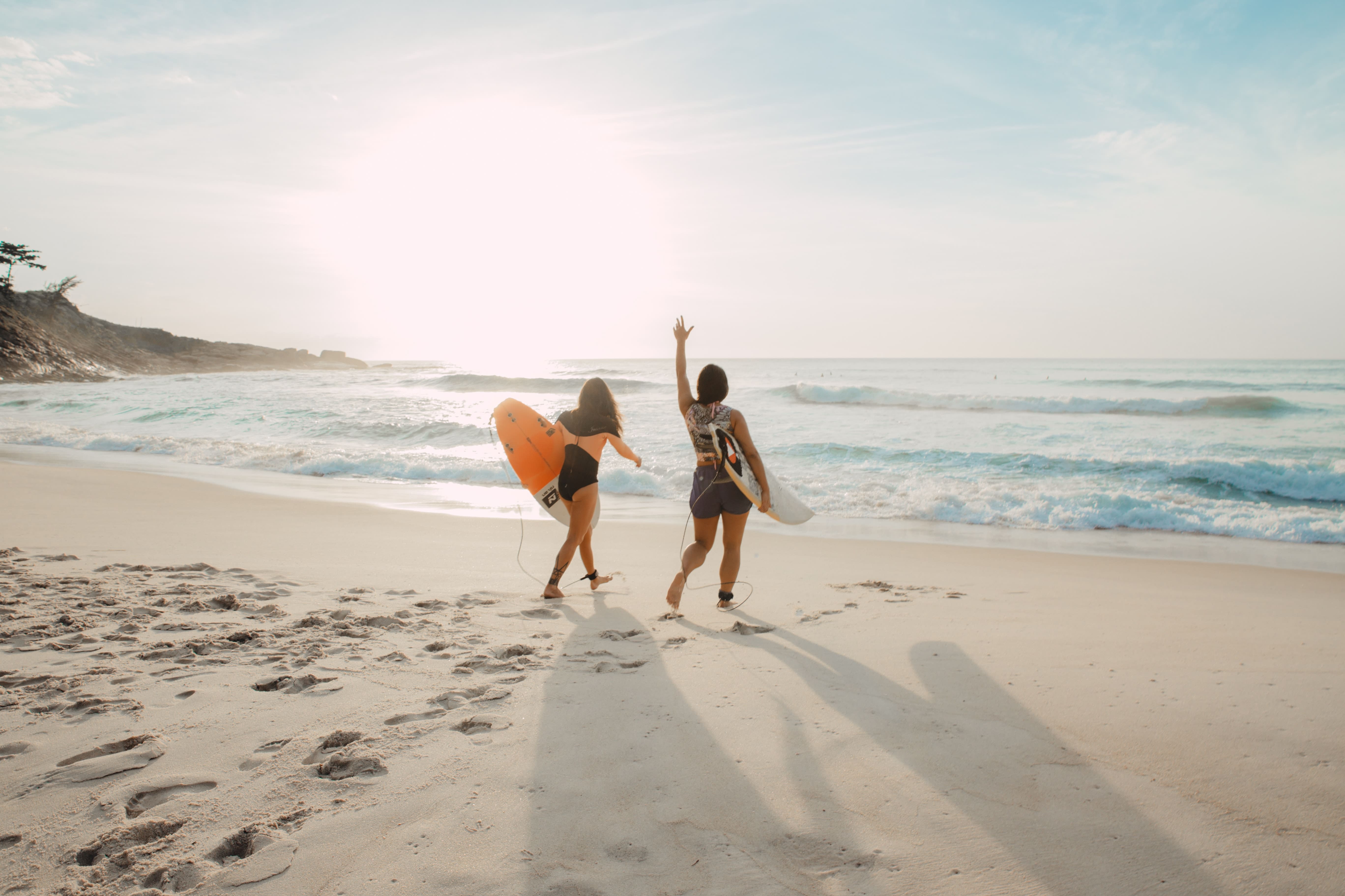 Zwei junge Frauen mit Surfboards laufen am Strand in Richtung Meer bei Sonnenuntergang. Symbol für Freiheit, Reisen und ferne Destinationen. 