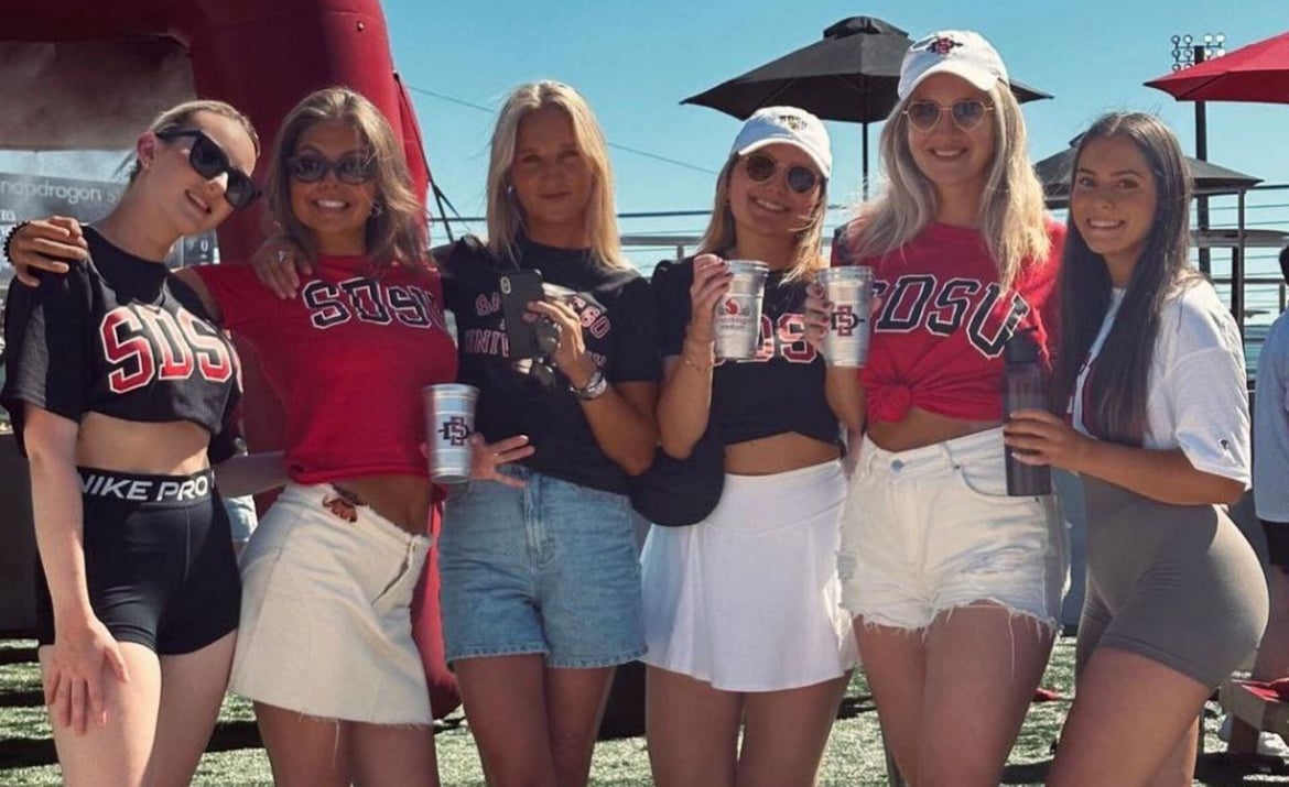 Sechs junge Frauen in SDSU-Outfits posieren fröhlich auf einem Sportplatz.