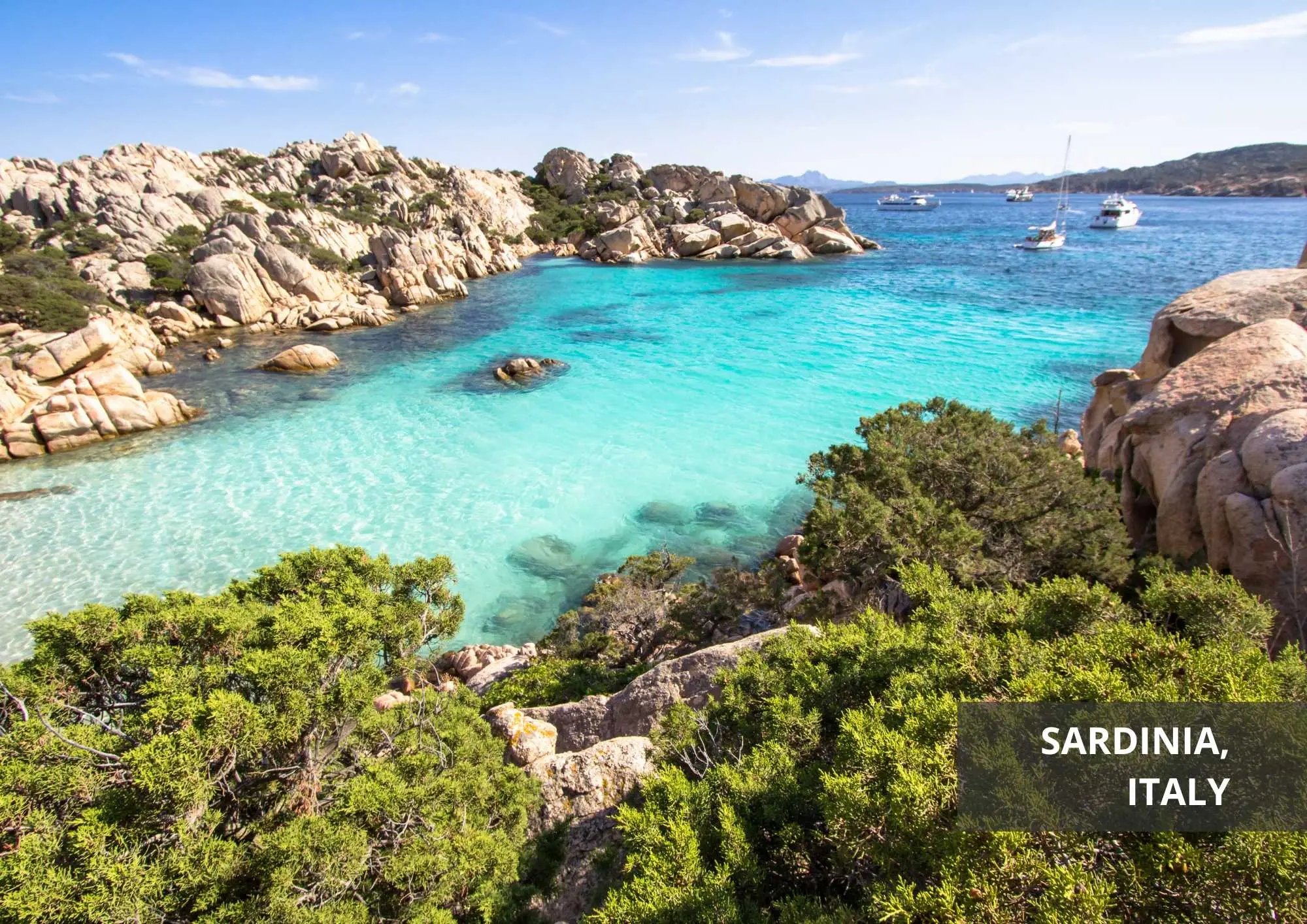 Traumhafte Bucht auf Sardinien, Italien, mit kristallklarem, türkisblauem Wasser, umgeben von felsigen Landschaften und grünen Büschen. Im Hintergrund ankern mehrere Segelboote im Meer.