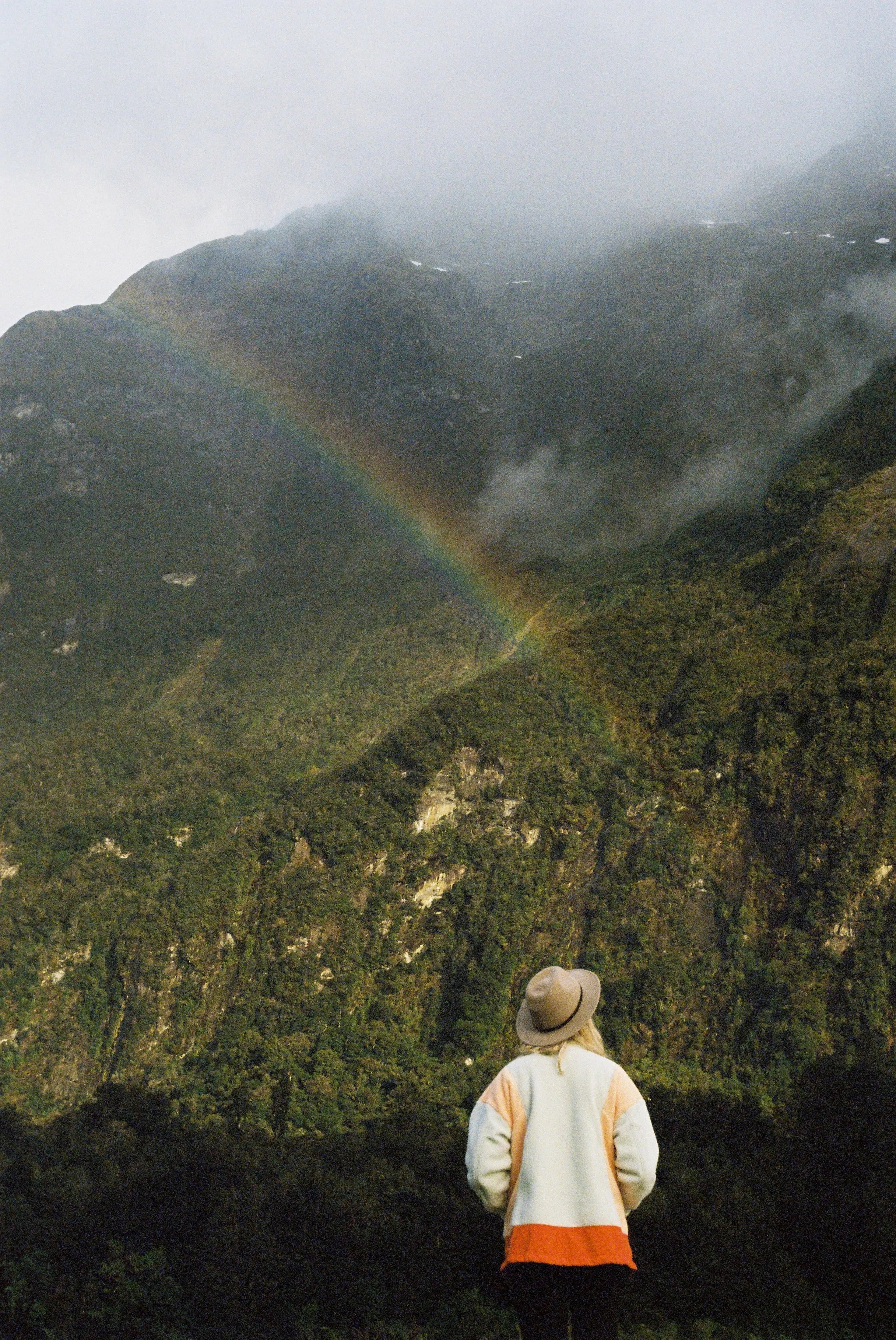 Person mit Hut blickt auf einen Berg mit Regenbogen – Symbol für Abenteuer und Naturerlebnisse.