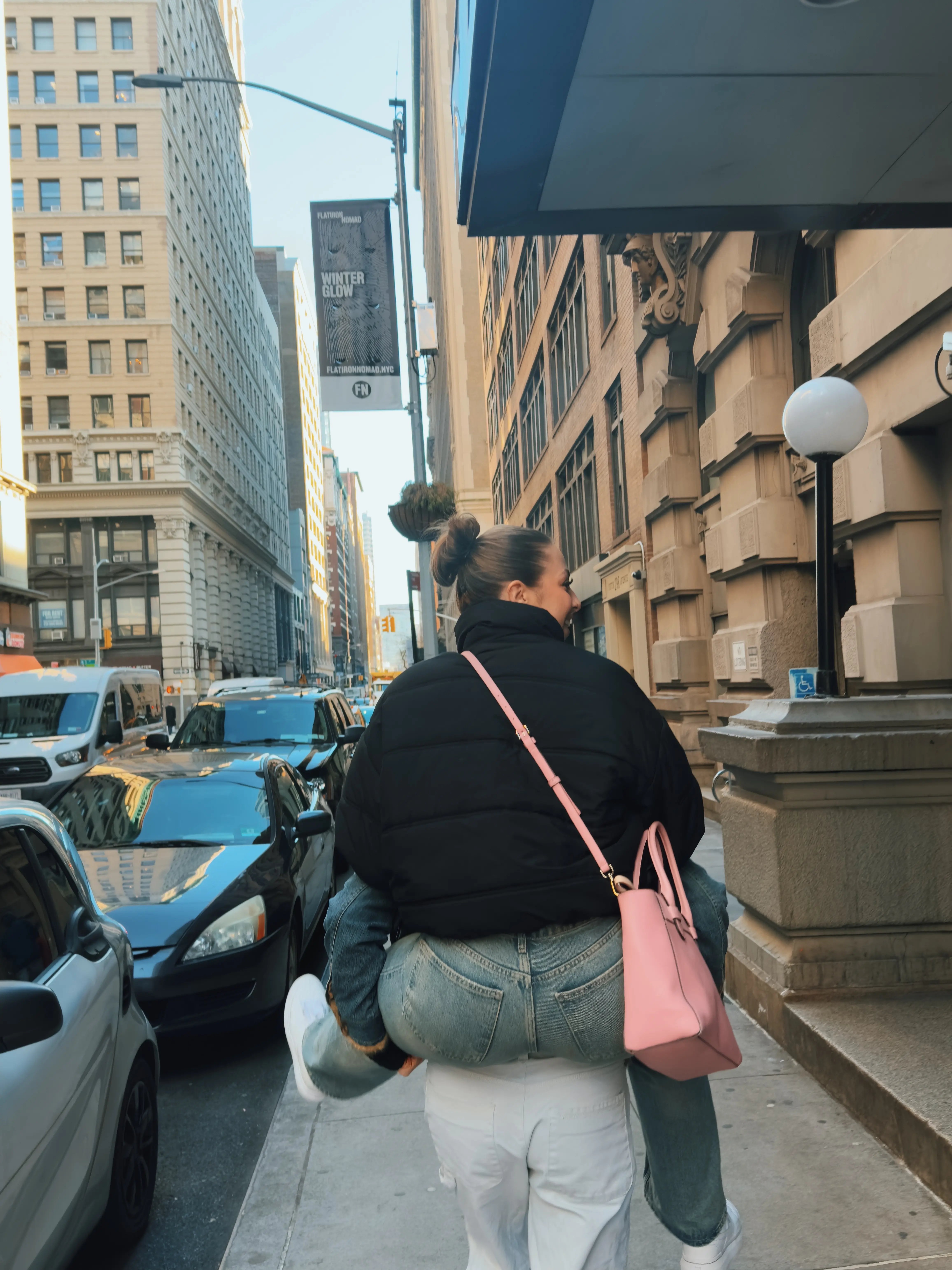 Zwei Personen in der City von hinten fotografiert, eine nimmt die andere huckepack, daneben viel befahren Straße.