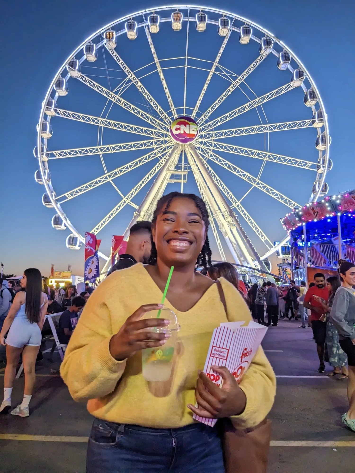 Junge Frau mit Getränk und Popcorn lächelnd vor einem beleuchteten Riesenrad auf einem Jahrmarkt.